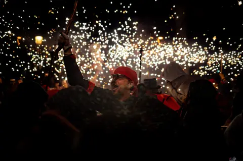 Matias Alvarez A man in a red bandana smiles and holds his arm aloft. He is part of a crowd of people celebrating and there are flying sparks in the night sky.