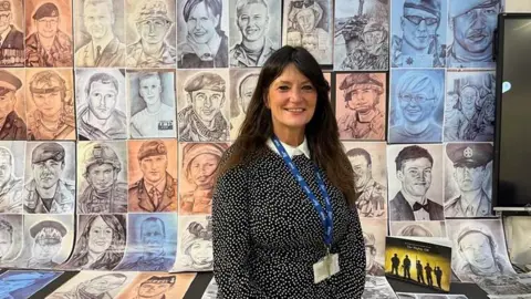 Artist Sam Bailey standing in front of a wall filled with artistic portraits of various military personnel. She is smiling, has long brown hair and is wearing a black dress with a white dotted pattern.