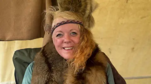 A woman with blonde hair, wearing a blue top with a fur scarf. She is also wearing a headband. 