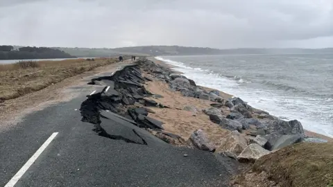 The A379 Slapton Line between Torcross and Slapton. The road is damaged with one lane washed away. The sea is to the right and there is a grassy patch and a lake to the left.