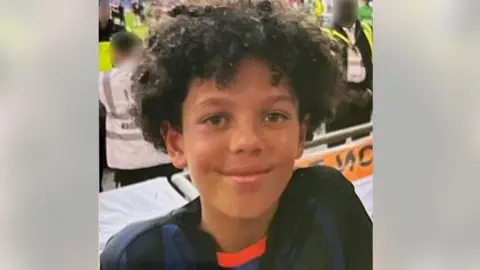 A boy is seen smiling at the camera. He is wearing a football jersey and there are people in the background at a football match