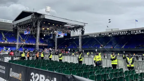 Connor Bennett/BBC View from the left side of the boxing arena from behind rows of chairs. Security in yellow hi-vis jackets are lined-up outside of the barriers next to the boxing ring.