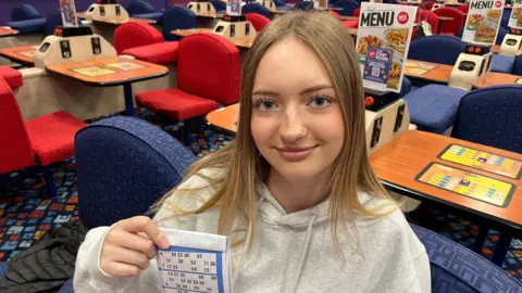 John Devine/BBC Alesksa Svencika has shoulder length blonde/sandy hair with a centre parting at the top of her forehead. She has a nose stud in her left nostril and is wearing a white hoodie type top and is holding a bingo card, behind her is a bingo hall with blue and red sofa style chairs with wooden tables for marking bingo cards.
