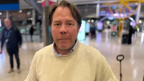 Alex Anderson has short dark hair and is wearing a cream jumper and blue shirt.