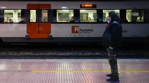 A man waits for a train at a station in Barcelona. A Rodalies train is seen on the opposite platform. Photo: 26 January 2026