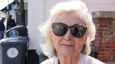 John Fairhall/BBC A head and shoulders image of a woman wearing a grey jumper and sunglasses. She is looking into the camera.