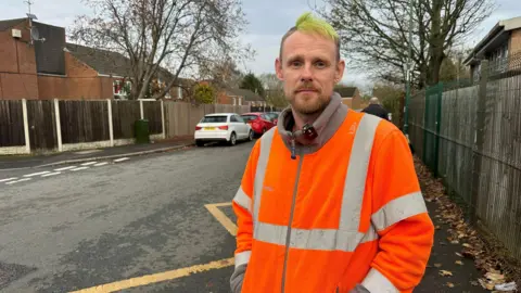 A man wearing a high vis jacket. He has green hair. He is stood in front of a road with cars parked along it.