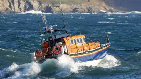 The existing lifeboat, which is a small blue and orange vessel, as it sails in rough weather, with the rugged Manx coastline in the background.