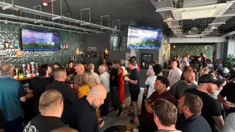 Members of the public standing by the bar, watching football on large TVs.