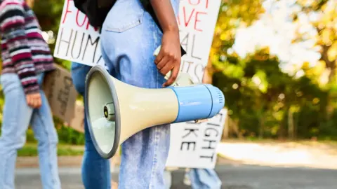 An anonymous person holds a megaphone down by their legs wearing blue jeans, with people holding protest placards in the background