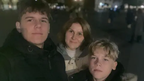 Ukraine Appeal The picture shows a woman, Iryna Maracine, pictured inbetween her 12 and 16 year old sons. It is night time, they are outside and wear winter coats.