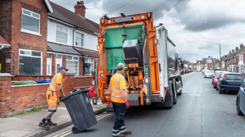 A bin lorry stops on a residential street with two bin men loading black bins into the back of the lorry before returning them to the kerbside