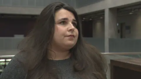 Nicola Love has long dark hair and is wearing a grey jersey. She is standing indoors in BBC Scotland's Pacific Quay building.
