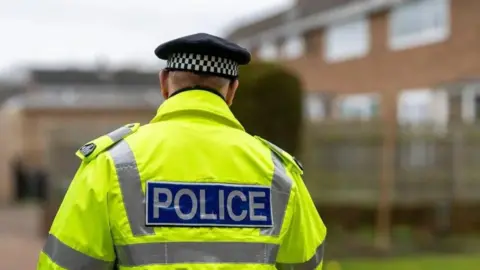 Getty Images A male police officer walking down a street with his back to the camera. He is wearing a black police cap and a luminous jacket.