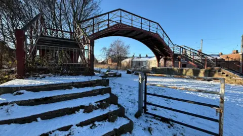 Jim Scott/BBC Snow covering an arched bridge which crosses a road. The ground is covered in snow with some footprints visible. Houses can be seen in the background with snow-covered roofs.