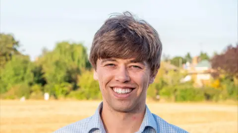 Cameron Holloway stares at the camera. He wears a blue and white checked shirt. In the background is a yellow field with green trees and hedgerows in the background. 