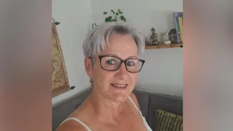 Mandy Hayward A woman with gray hair and glasses. The head and shoulder shot shows Mandy smiling. Behind her is a grey sofa and a shelf with various trinkets on such as a Buddha a candle and some books.