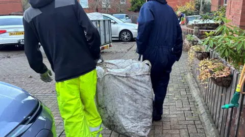 A man in a navy boiler suit holds a large white sack with his left hand. On the other side of the bag is a man wearing yellow hi-viz trousers and a black hoodie. They walk on a grey brick drive way, with their back towards the camera. You cannot see either of their heads. There are a group of several different cars in the background. 