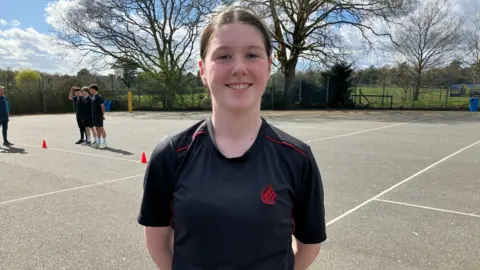 Leanne Rinne/BBC Caitlin is stood in the school playground in a school branded black top with red piping and a red logo. She has her hair tied back and her hands behind her back.