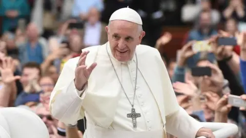 PA Media The late Pope Francis, an elderly man wearing a white skullcap-style hat over grey hair, with white robes and a large silver crucifix hanging around his neck. He is smiling and waving as crowds of people behind him take photographs.