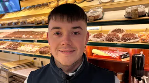 A young man smiles for a photograph in a bakery. He wears a dark gilet and light coloured top underneath and behind him are a variety of cakes. 