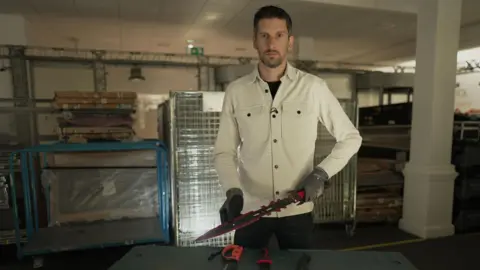 BBC BBC News's Dan Johnson holding one of the purchased knives. He is holding the large knife in two hands while wearing gloves, looking directly to camera