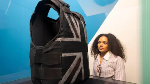 David Parry/PA Media A woman looking at a black and white union jack stab vest which sits on display. The woman is wearing a white and light pink striped blouse with her glasses resting on the neck line. 
She has curly dark hair and a nose stud. 
The background is the clean lines, coloured blue, white and green of an art gallery.