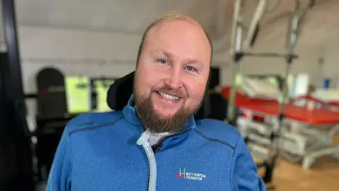 Matt Hampson is sat in his wheelchair in a hall at the Matt Hampson Foundation, with various rehabilitation equipment in the background. He is smiling and wearing a blue fleece with the Foundation's logo on it.
