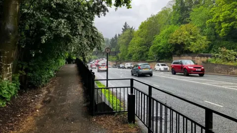 Footpath next to a dual carriageway which has cars on both sides of the road and a queue of traffic forming ahead. There are black metal railings between the path and the road. There are trees and dense foliage on both sides of the road.