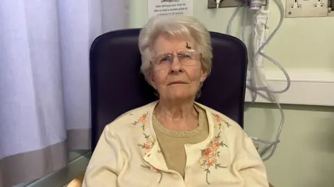 Joan Elkington, who is an elderly woman dressed in a cream cardigan with pink and yellow flowers on it. She is wearing glasses and sitting in a chair in the corner of a hospital room.