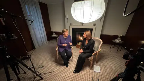 BBC Former German Chancellor Angela Merkel sits talking about her time in office with the BBC's Katya Adler in a hotel room. Their chairs are facing each other and there is a variety of broadcast equipment in view