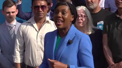 PA Media Kemi Badenoch speaking, with supporters stood behind her. She is wearing a green top and light blue blazer.