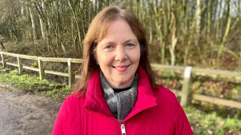 Claire Ward, wearing a red puffer jacket and grey scarf, smiles for the camera. Behind her there is a path, a wooden fence and some trees.