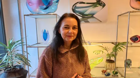 Nicola Williams who owns Kinki Glass is standing smiling into the camera with some glass art work sitting behind her on shelves 