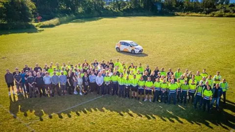 A group of people from above, many in high visibility tops, stand in a field with a Sure branded car behind them.