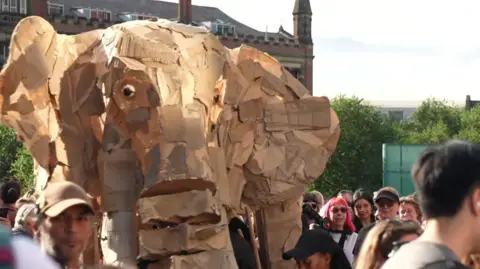 A crowd of people are towered over by a wooden elephant puppet 