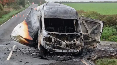 Dorset and Wiltshire Fire and Rescue Service The van is parked at the side of the road. Much of it is black and clearly wrecked by the fire. A once white panel has been seemingly blown open by the fireworks on the left side of it, while the right seemed to have detached. 