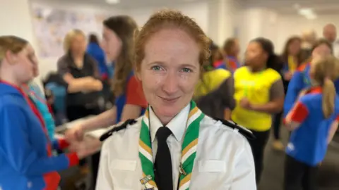 A ginger-haired woman in a police uniform looks at the camera with girls milling about in the background