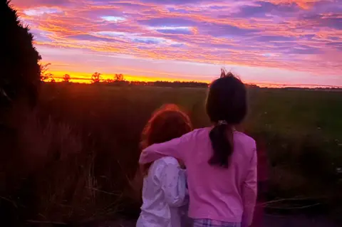 Lori Carnochan Two children in pink pyjamas watching a sunset. The sky is pink, purple and yellow. 