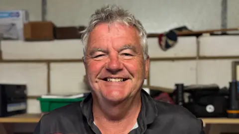 A head and shoulders image of Michael Hancock. He is wearing a black and red top and standing in front of a shelf that has various items on it. He is smiling and looking directly into the camera. 
