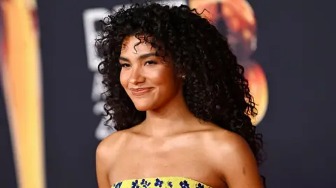 Olivia Dean is wearing a yellow strapless dress with navy blue flowers. Her hair is dark brown and curly, she has brown eyes. She is smiling slightly to the left of the camera. 