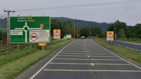 Google A629 dual carriageway which separates the villages of Steeton and Silsden in Bradford. Fields to the left - hills beyond, road sign with roundabout ahead, road with yellow lines to slow for roundabout, telegraph wires across the road.