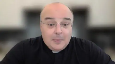 Fr Domenico is speaking on a Teams call. He wears glasses and a priests' clothing. The background behind him is blurred.