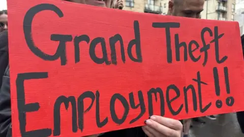Banner from protestors outside Rockstar Games' offices reading 'Grand Theft Employment!' It's in black writing on a red background being held by what appears to be a woman's hand