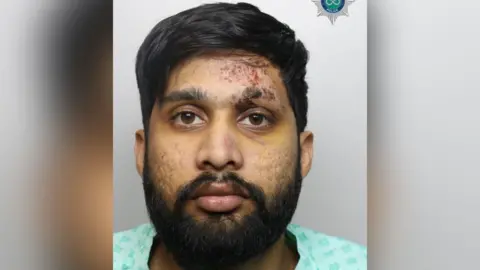 Staffordshire Police A mugshot of a man with dark hair and a dark beard wearing a light blue shirt.