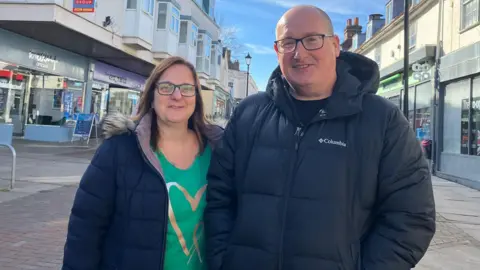 Josh Holmes-Bright/BBC A women wearing a blue coat and green t-shirt standing next to a man in a blue Columbia coat on Harlow high street