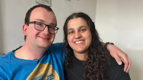 A man in a blue t-shirt and black glasses has his arm around his wife. The women has dark curly hair and is wearing a dark blue jumper and jeans. They're sat on the sofa in their living room.