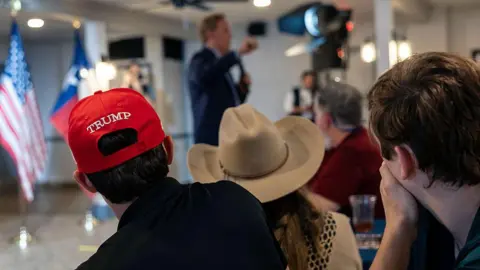 Getty Images Attendees of a Bill Paxton rally watch him speak. Two of them wear hats; one of which is a signature red Trump cap