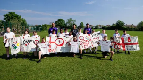 Rogan Thomas A group of school pupils at Headley Park Primary School hold up several banners showing their support for Bristol City ahead of their game against Preston. Standing behind the children, who are on the school's playing field, are City players Zak Vyner, Joe Williams and Elijah Morrison.