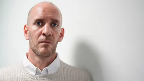 PA Media Dan Barr, 41, who has blue eyes, a bald head and light stubble, speaks into the camera wearing a beige jumper over a white shirt. 